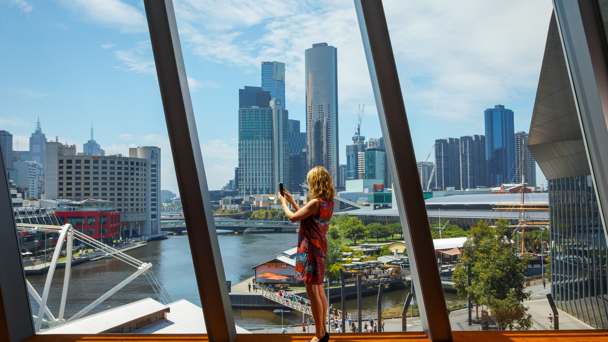 Woman taking photo at the floor to ceiling window from inside MCEC.  The view is the city landscape, greenery, buildings, outside South Warf precinct 