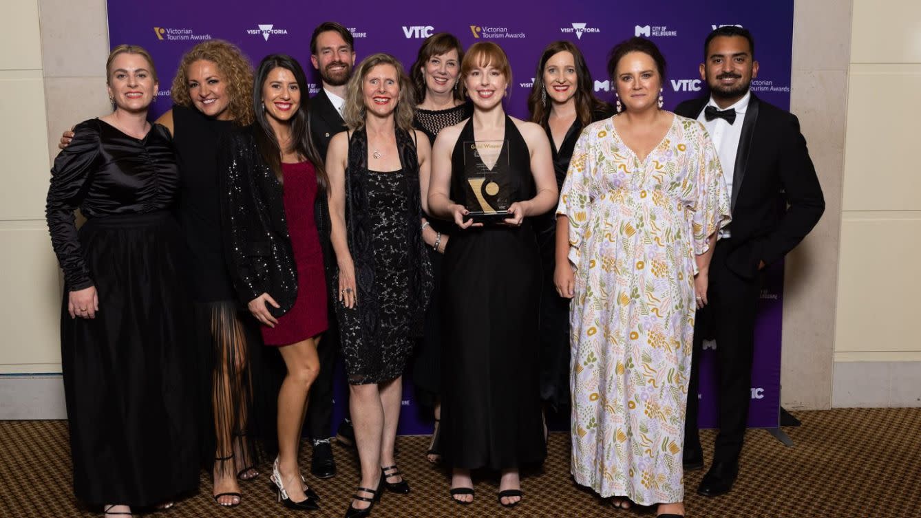 Group of people in formal black tie attire, one woman in the middle holds a trophy. They are all standing in front of a purple back drop which has the logos of Visit Victoria, City of Melbourne and VTIC across it. 