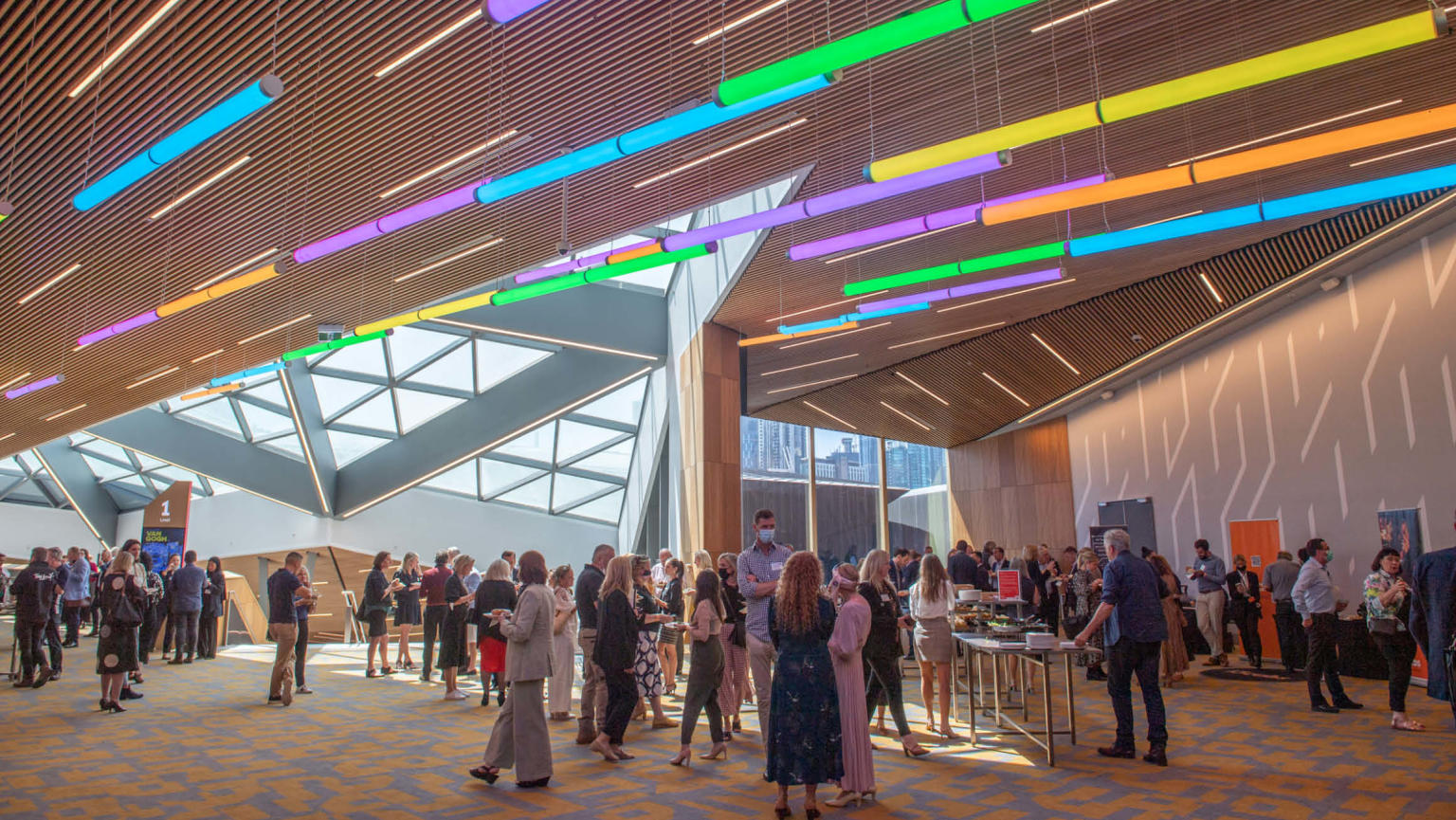 A lively foyer filled with a diverse crowd engaged in conversation and mingling. Overhead, vibrant bars of multi-coloured lights in shades of blue, purple, green, yellow, and orange hang from the ceiling.