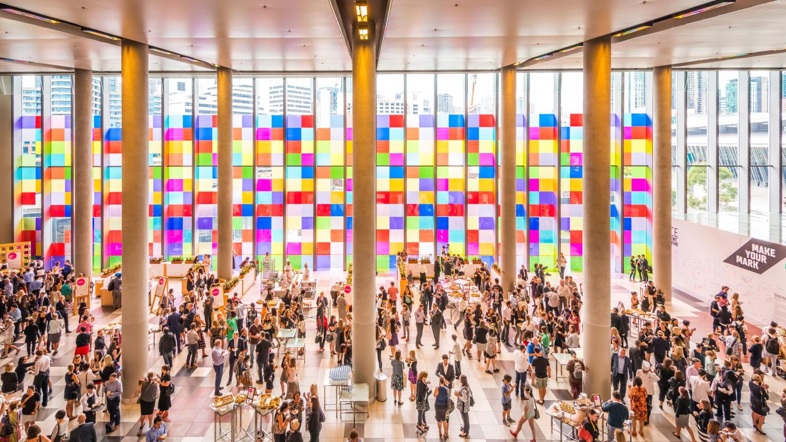 Overview of a crowd in the Convention Centre Foyer. Colour squares cover the floor to ceiling windows with light streaming through. 
