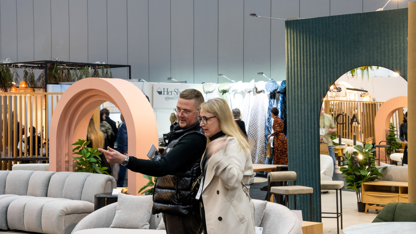 A couple of people point to something off screen. A furniture display in an exhibition building is seen behind them. 