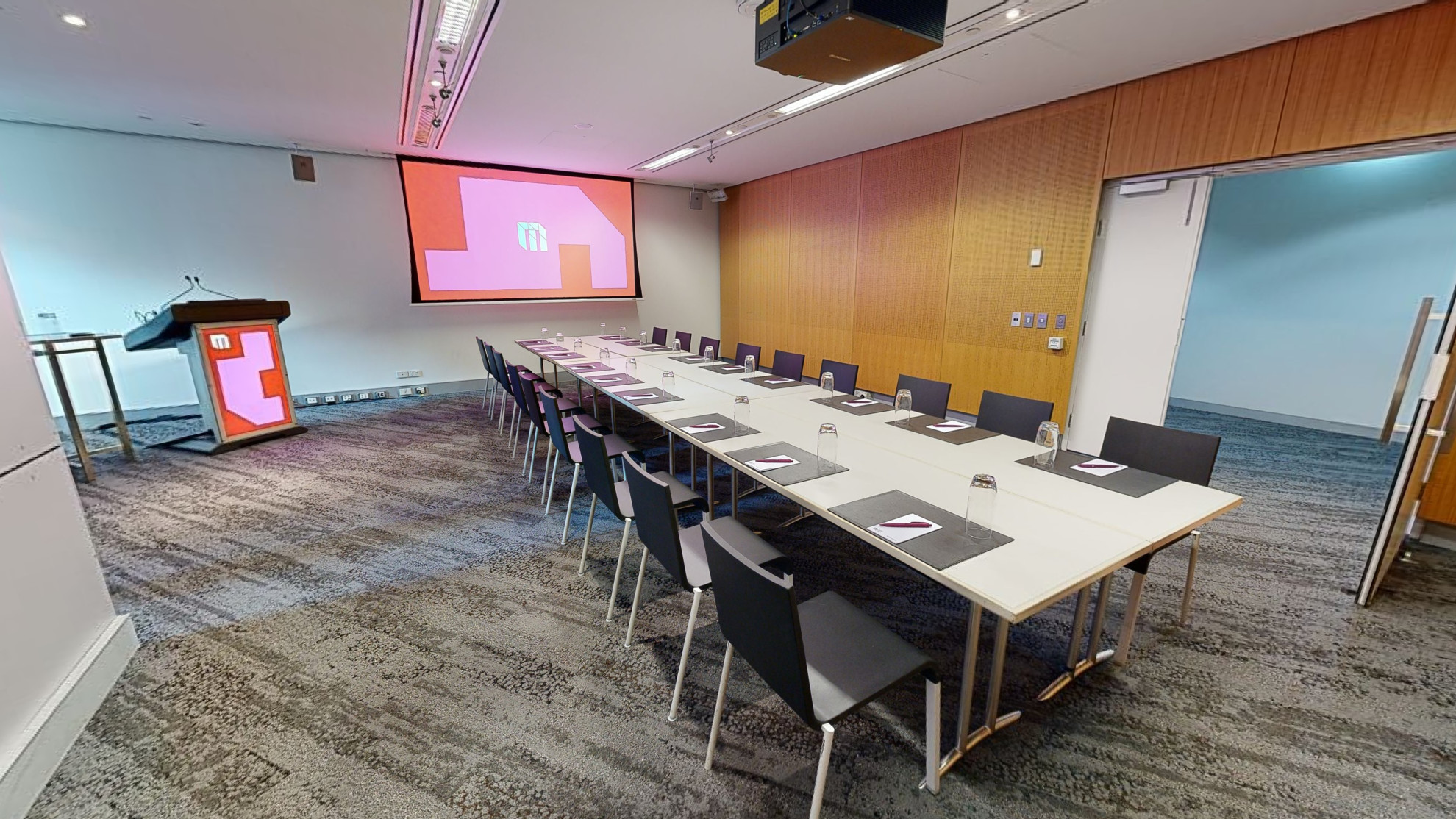 A conference or meeting room featuring a spacious boardroom layout equipped with a sizable screen and comfortable chairs for attendees. A lectern also sits in the corner to the left of the screen. The double doors to the room are open in the right corner of the image. 