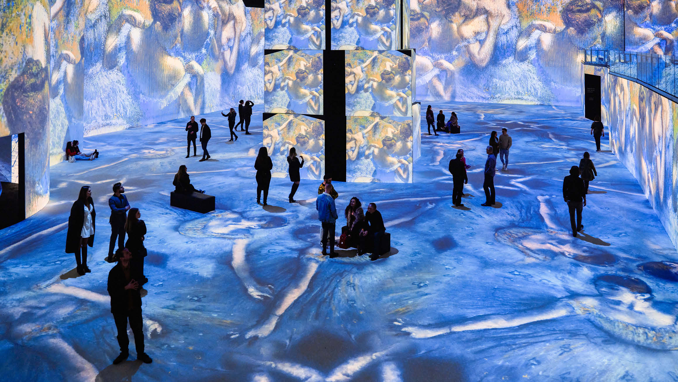 An image capturing individuals immersed in a transformative gallery experience at THE LUME Melbourne. Surrounded by expansive digital screens, they are engrossed in the Monet and Friends Exhibition, this particular image showcases Edgar Degas' blue ballerina paintings.