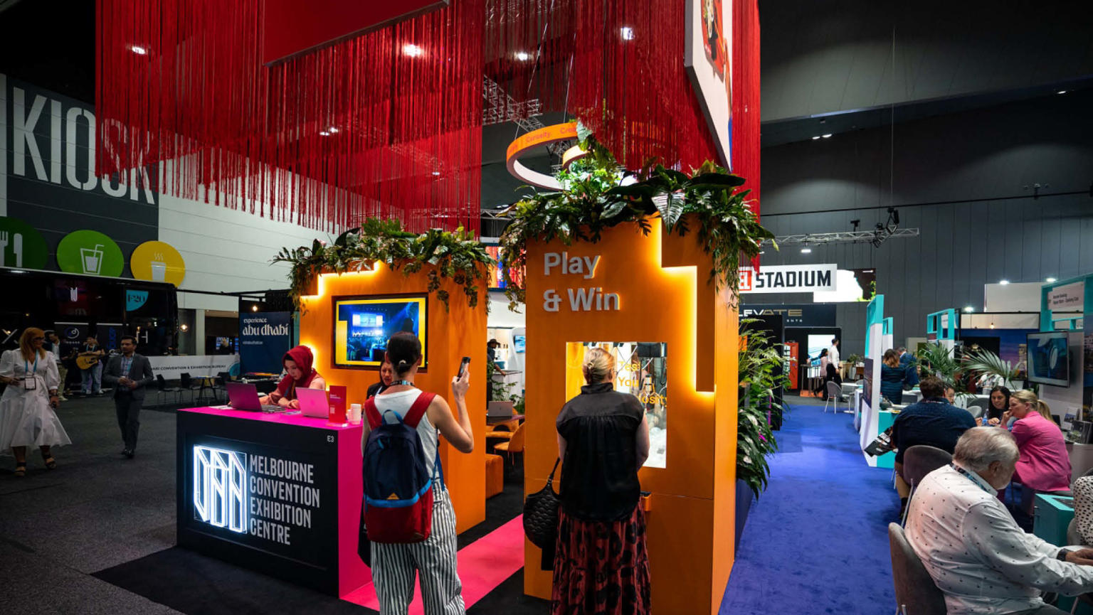 An attention-grabbing exhibition stand with a dynamic arrangement of elements: long red fringing hanging from the ceiling, a vibrant orange wall featuring a walk-through cutout. On the left side, a pink and black table proudly displays a neon logo reading 'Melbourne Convention and Exhibition Centre.' To the right, a captivating claw drop game beckons with the enticing words 'Play & Win' above it. Excited individuals are gathered around the stand, adding to the lively atmosphere.