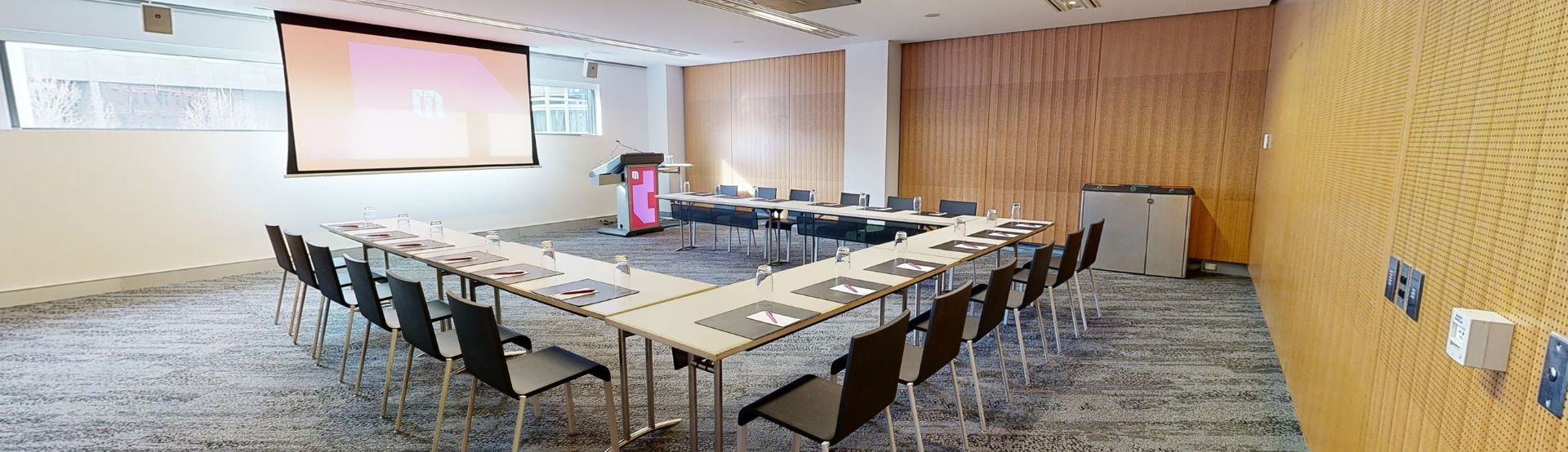 A meeting or conference room with tables and chairs arranged in a u-shaped boardroom configuration. The tables and chairs are facing a large projector screen and lectern both which have MCEC branding displayed on it. 