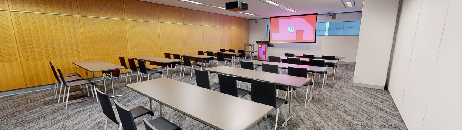 A meeting or conference room featuring neatly arranged tables and chairs in rows, providing a professional setting for meetings and discussions. The tables and chairs face a large projector screen and lectern at the front of the room. A small strip of windows that sit at eye level run behind the screen. 