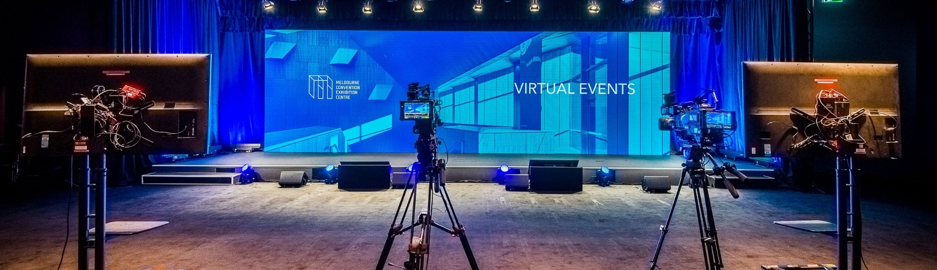 Two cameras and two screens are set up facing a large blue backdrop and stage. The MCEC logo and the words "Virtual Events" is displayed on the backdrop. 