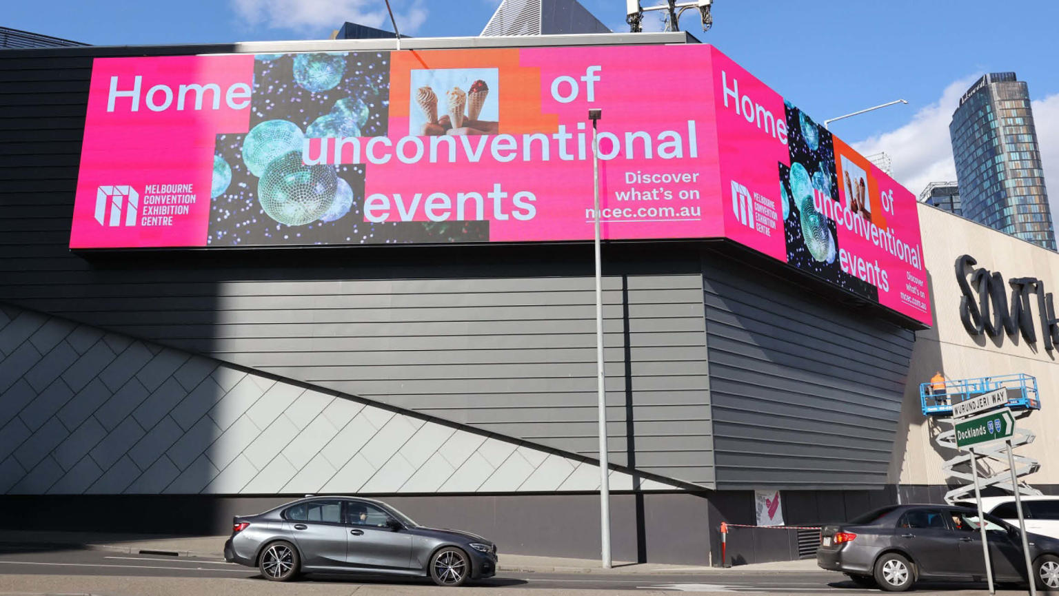 
A sizeable grey building exterior featuring a captivating digital sign in the foreground. The sign displays the vibrant message "Home of unconventional events" alongside imagery of disco balls and ice cream cones. The Melbourne Convention and Exhibition Centre logo is positioned in the bottom left corner of the sign. Cars can be seen driving past the building.