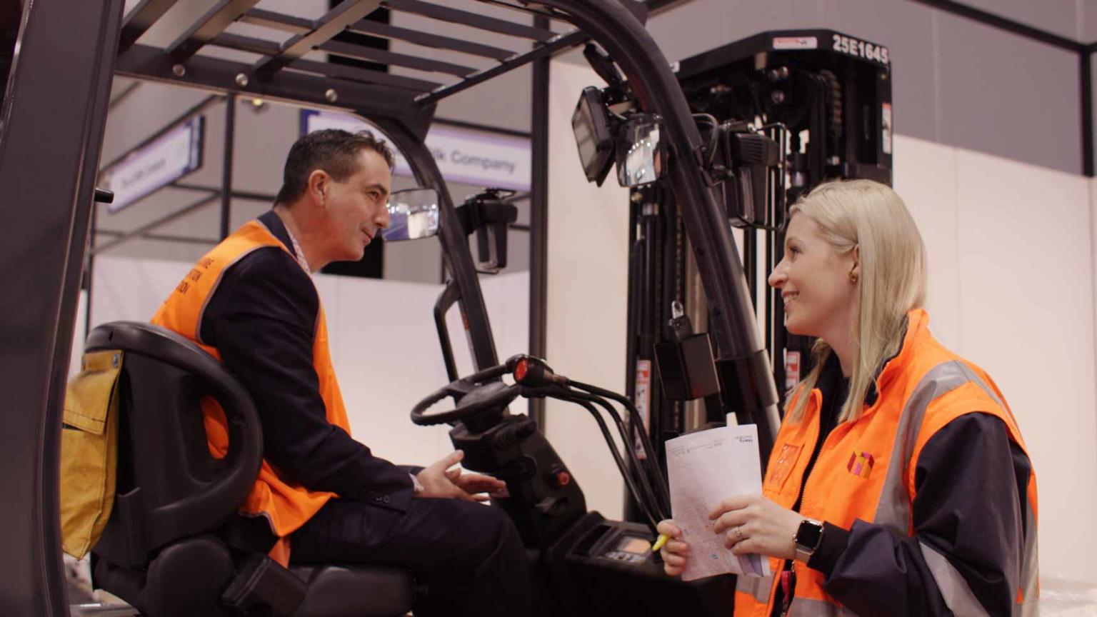A man on a forklift talks to a woman standing next to it. They both are wearing high visibility orange vests. 