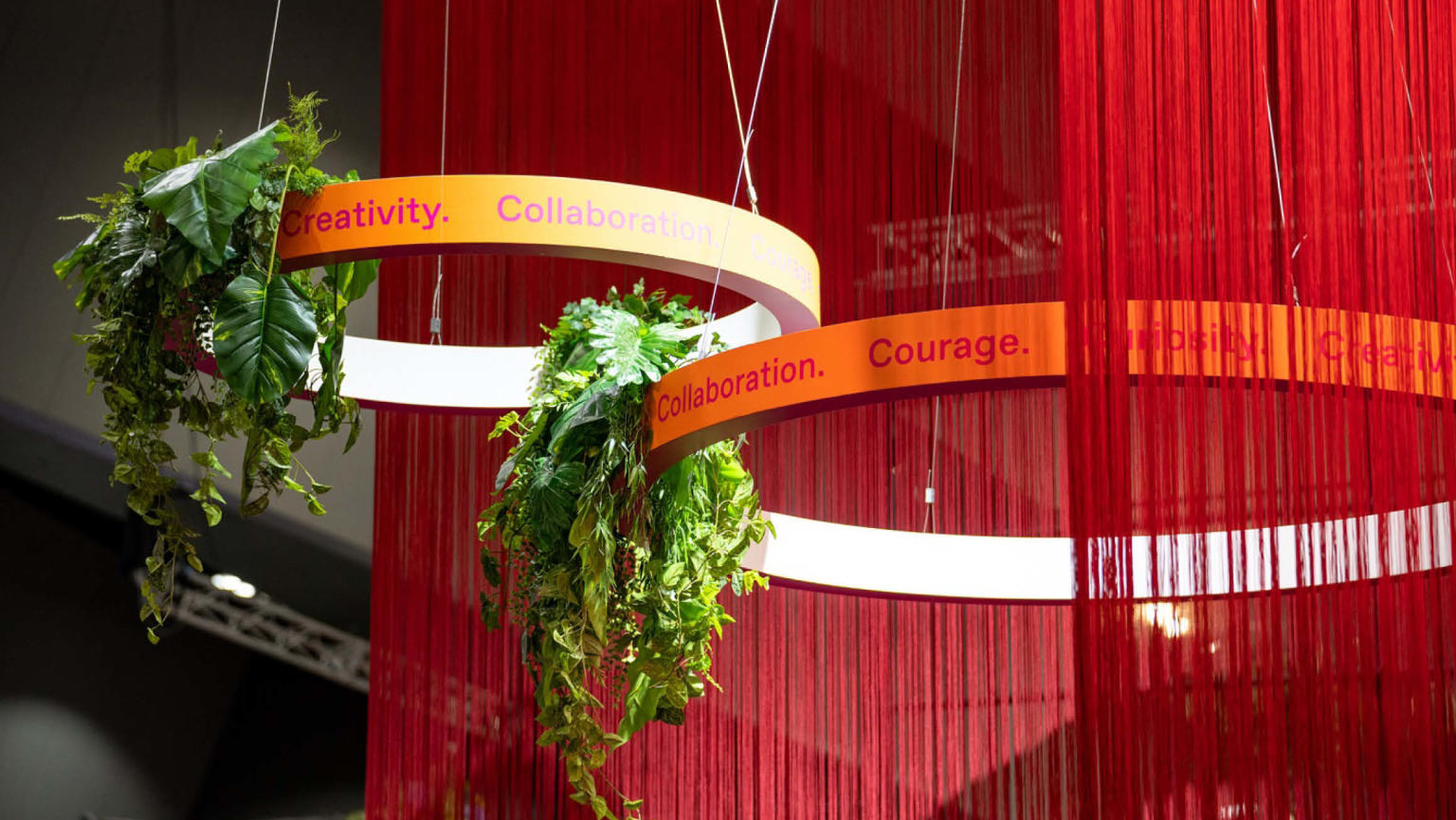 A close-up view of vibrant long red fringing with two hollow circle bands at the centre. The bands are adorned with the inspiring words 'Creativity, Collaboration, Courage, and Curiosity' and embellished with hanging greenery.