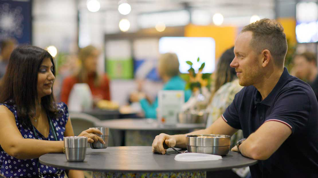 A man and woman sit at a table talking with stainless steel cups and bowls on the table. 