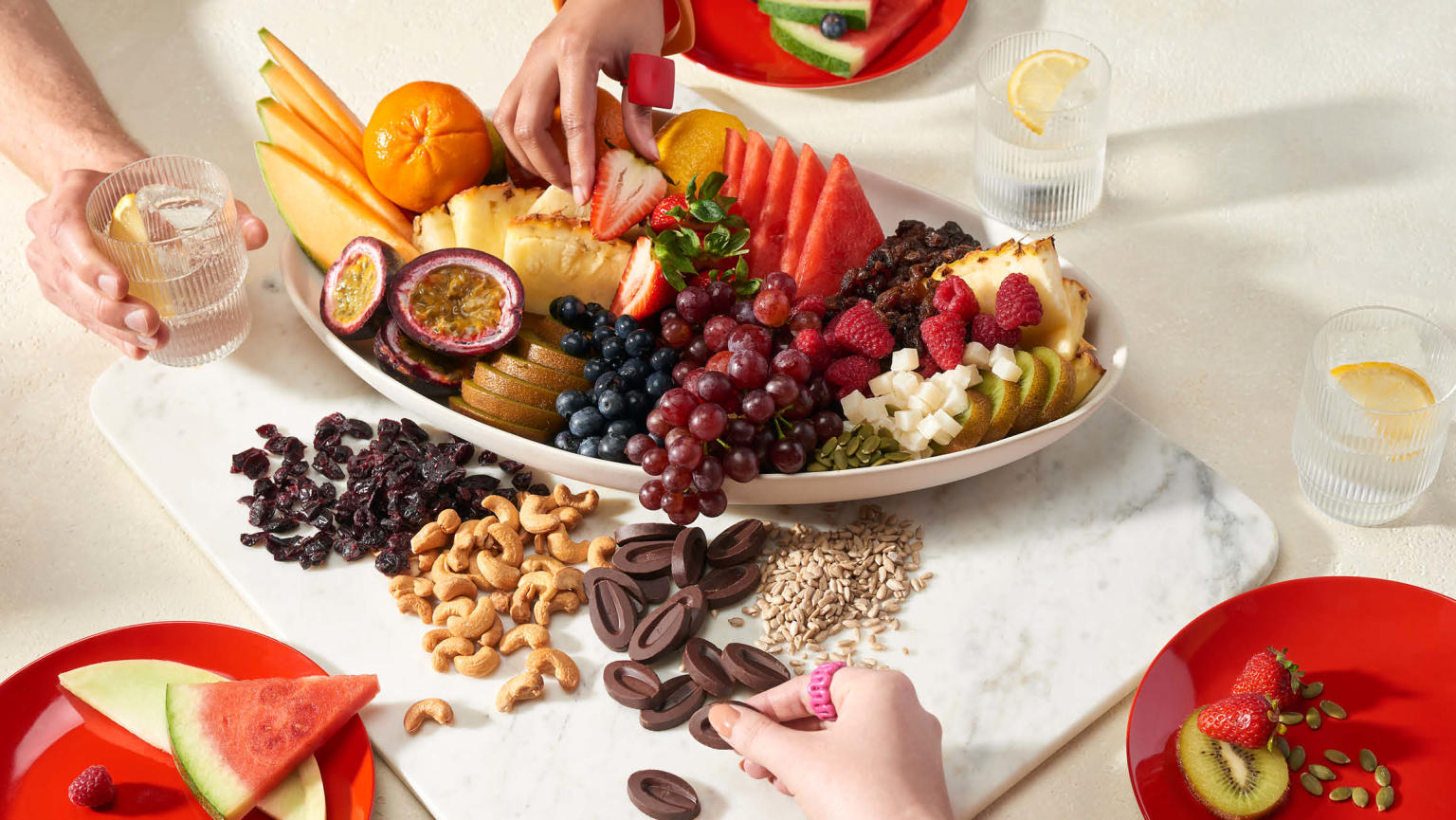 An appetising assortment of fruits and nuts arranged on platter on top of a stylish marble board on a table, surrounded by three red plates with fruit on them, with three hands reaching out to grab a variety of delectable items, including a refreshing drink. 