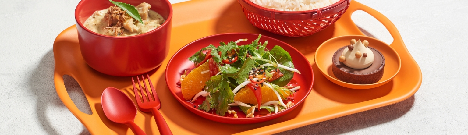 Four small dishes in red and orange tableware sit on top of a bright orange tray with a matching red spoon and fork lay to the side. 