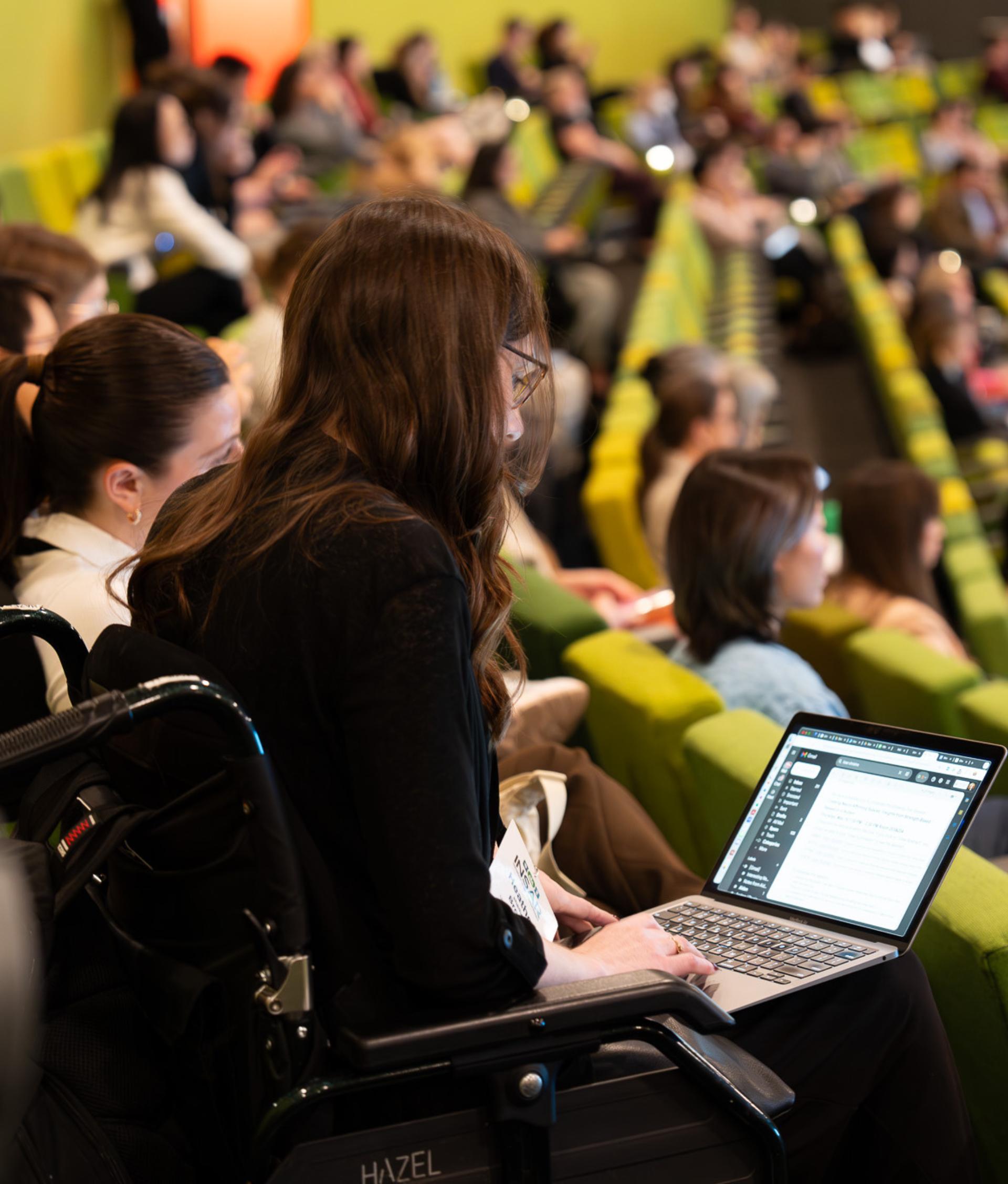 A person is sitting in a wheelchair at the end of a row of green seats in a theatre. The seats are filler with people. 