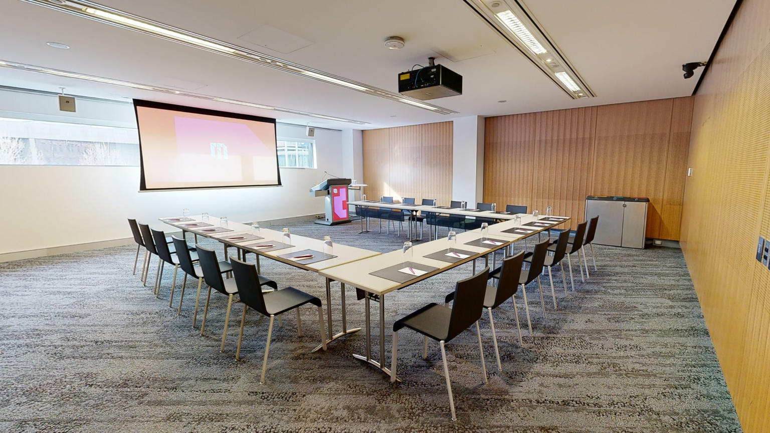 A meeting or conference room with tables and chairs arranged in a u-shaped boardroom configuration. The tables and chairs are facing a large projector screen and lectern both which have MCEC branding displayed on it. 