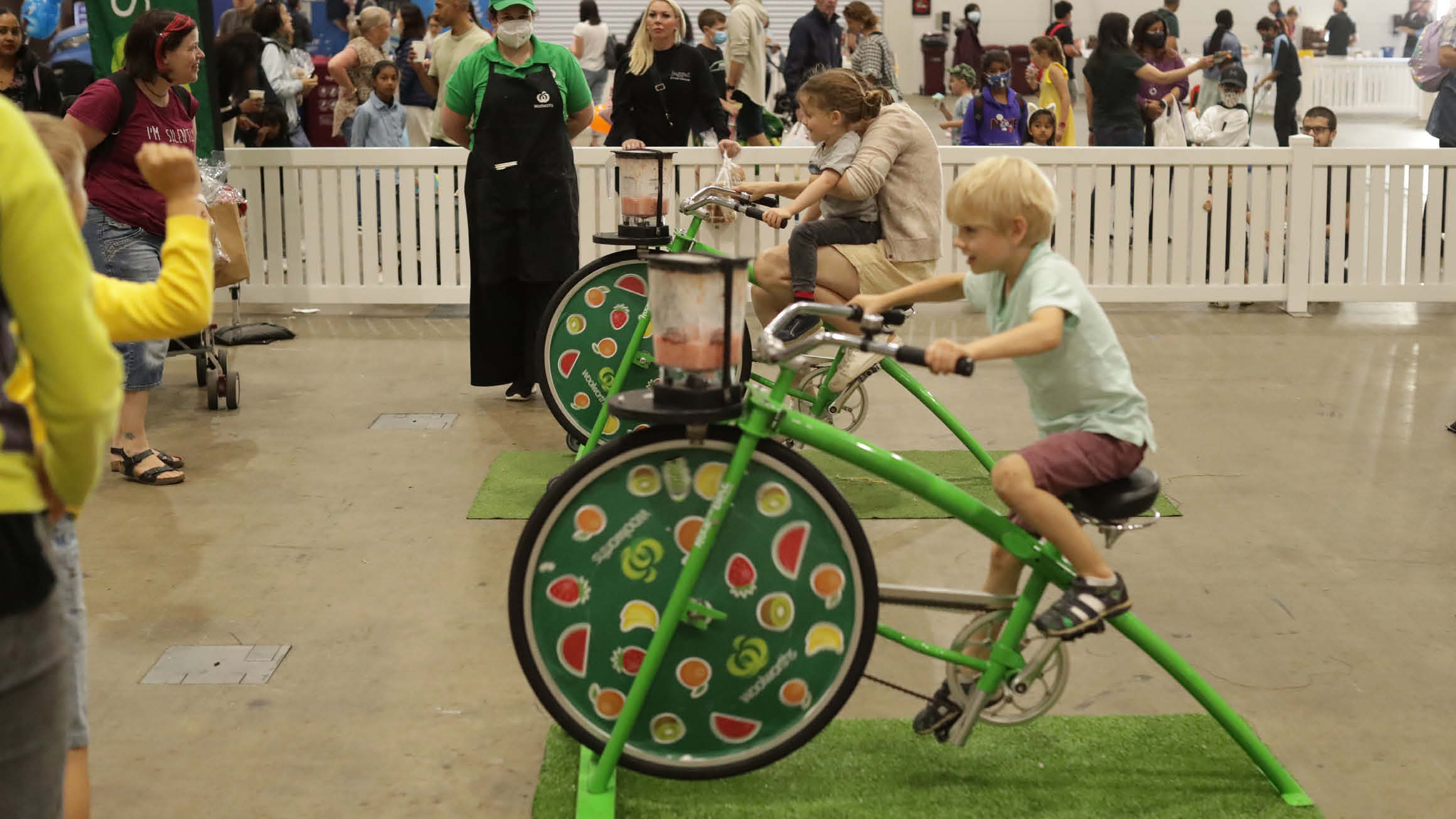 Two kids on stationary green bicycles that have vegetable stickers all over the front wheel. 