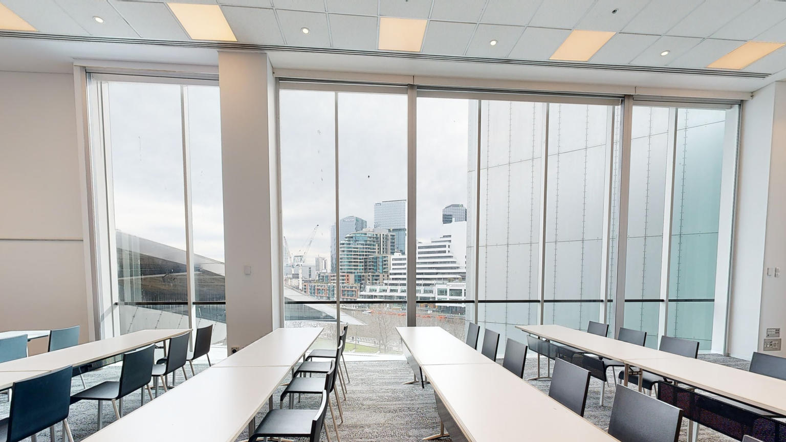 Table and chairs are set out in a classroom formation of rows facing the left side of the image. A large floor to ceiling window fills one wall of the room, which looks out to the Melbourne city. 