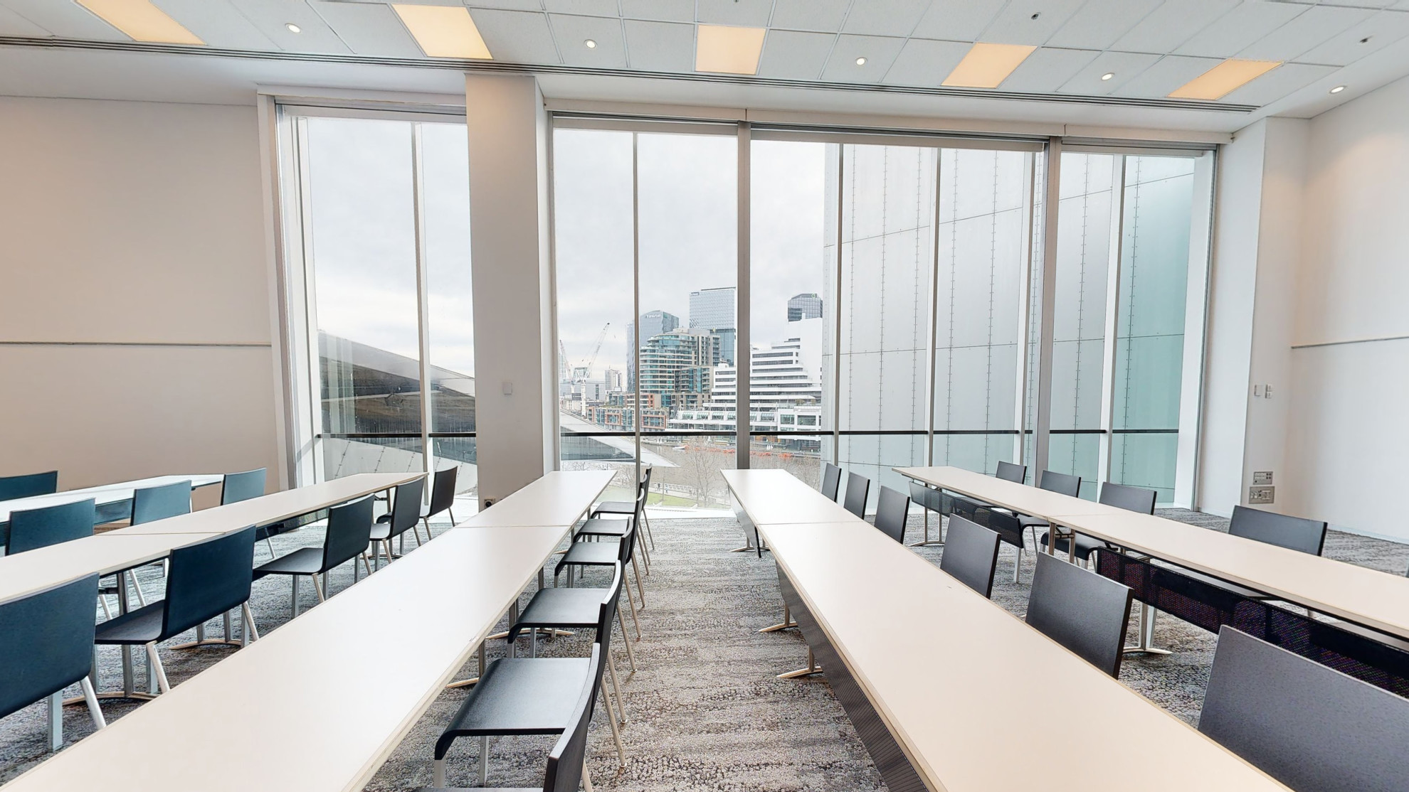 Table and chairs are set out in a classroom formation of rows facing the left side of the image. A large floor to ceiling window fills one wall of the room, which looks out to the Melbourne city. 