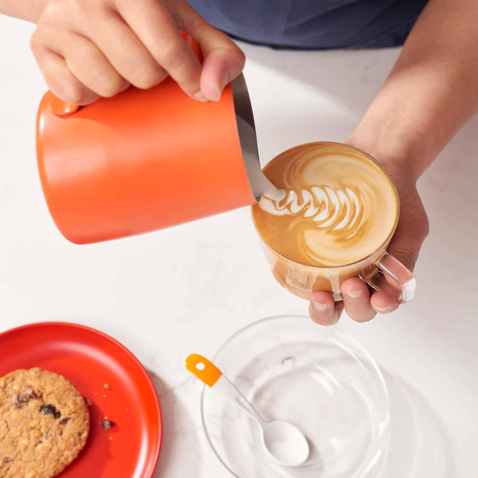 Two hands, one holding an orange milk jug pouring milk into a coffee cup being held by another hand. A red plate sits in the corner with a cookie on it. 
