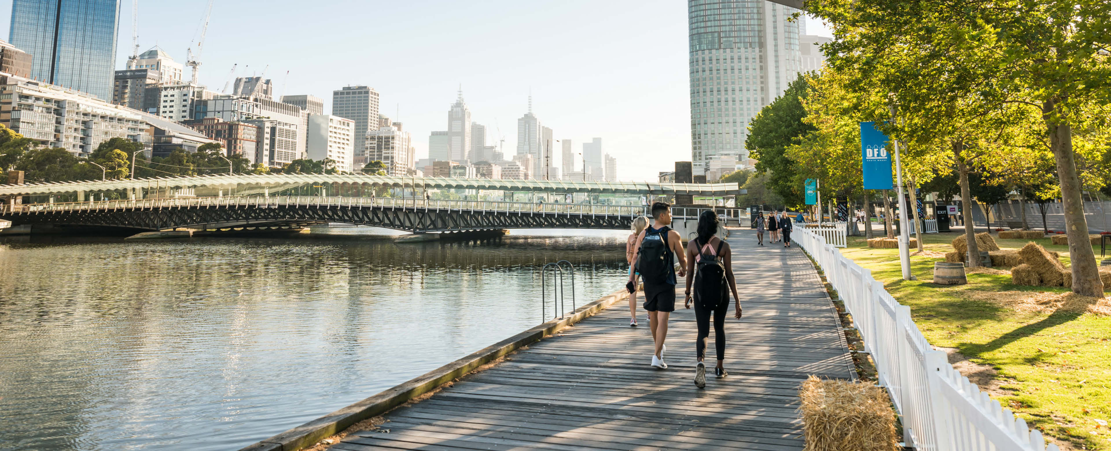 An image capturing the South Wharf precinct in Melbourne. A couple strolls along the footpath with the Yarra/Birrarung River on their left, while a line of trees is on their right side. In the distance, a bridge spans the river, with the city skyline in the background. 