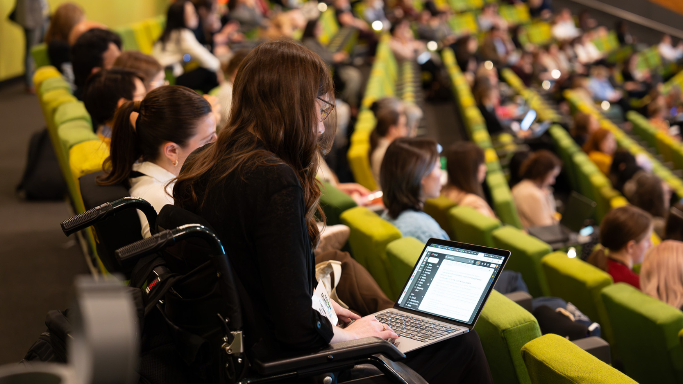 A person is sitting in a wheelchair at the end of a row of green seats in a theatre. The seats are filler with people. 