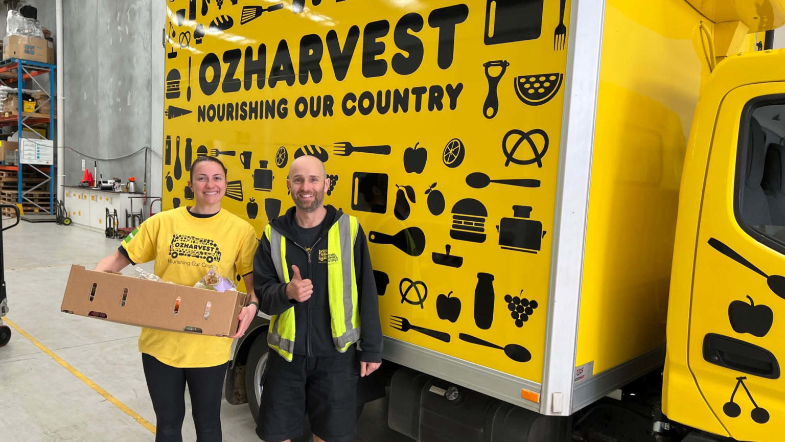 An image showing two people - one holding a cardboard box of food and wearing a yellow OzHarvest t-shirt, and the other wearing a black hoodie and a yellow high vis vest doing a thumbs up - standing and smiling in front of a yellow OzHarvest van. 