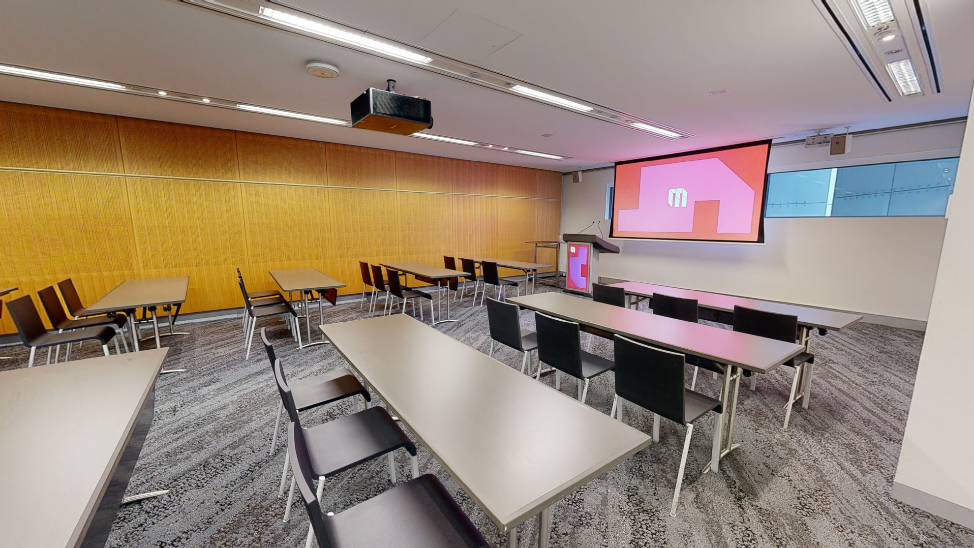 A meeting or conference room featuring neatly arranged tables and chairs in rows, providing a professional setting for meetings and discussions. The tables and chairs face a large projector screen and lectern at the front of the room. A small strip of windows that sit at eye level run behind the screen. 