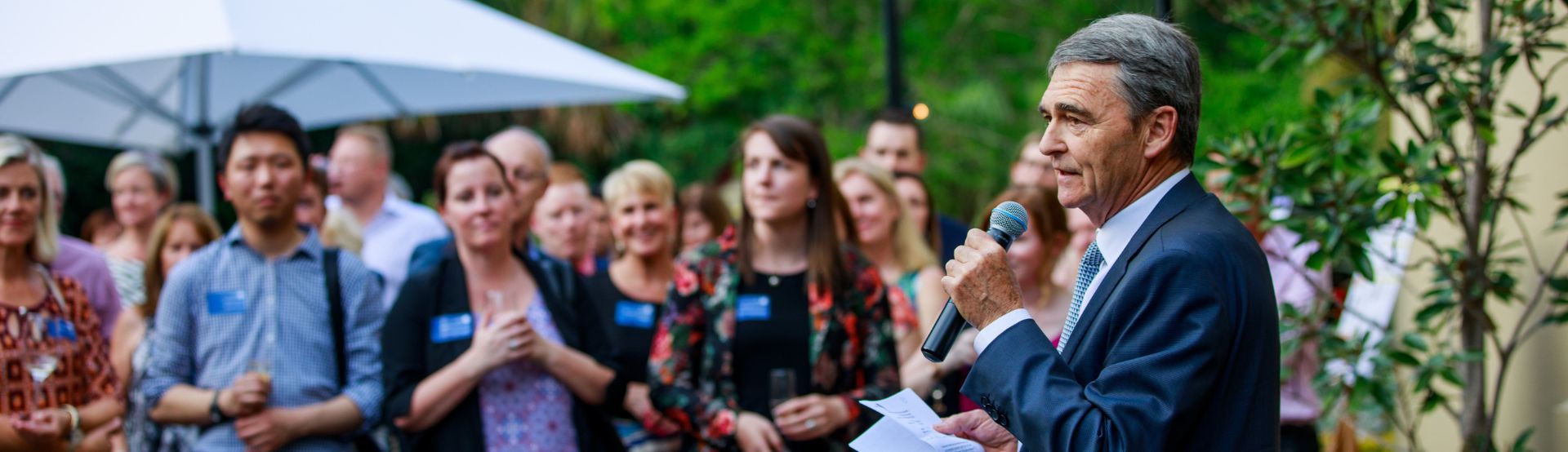 A crowd in the distance looks towards a man in the foreground wearing a suit, talking into a microphone. 