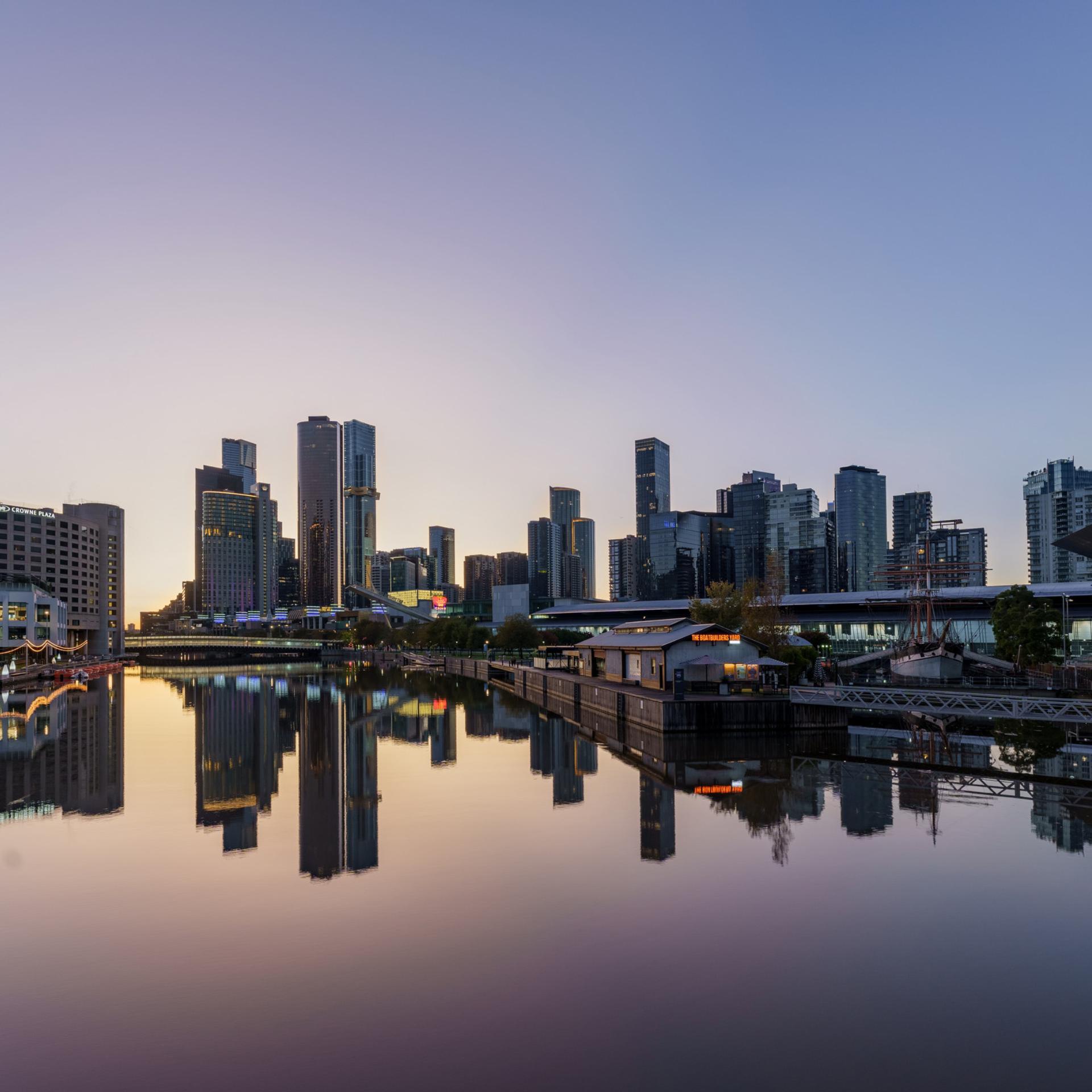 Outside view of the city along the Yarra River. MCEC can be seen to the right with tall skyscrapers behind. The sun is setting so the deep blue sky is reflected in the water. 