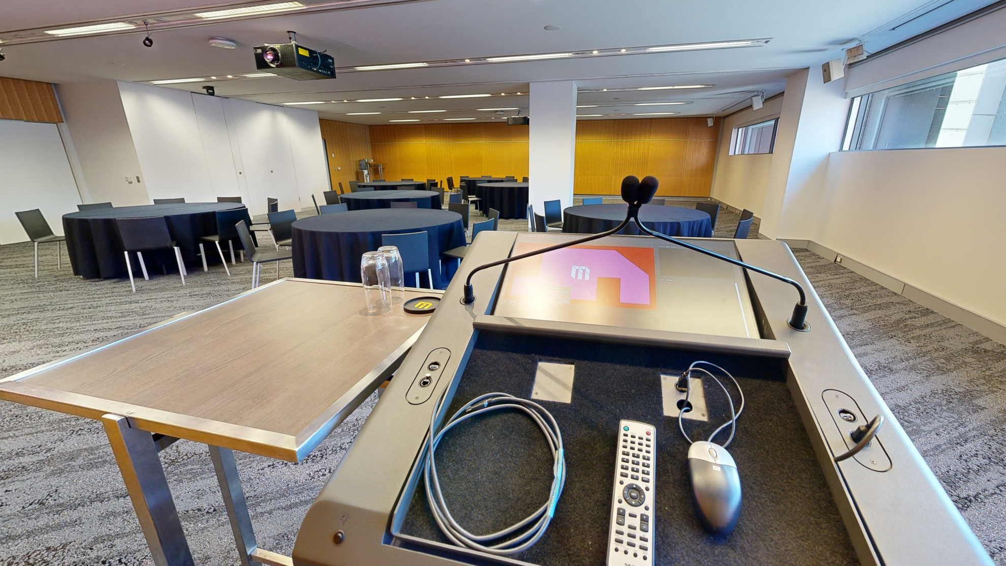 A conference or meeting room with arranged round tables and chairs for meetings or discussions. The tables focus around facing a lectern at the front of the room. A strip of windows that sit at eye level run along side one wall. 