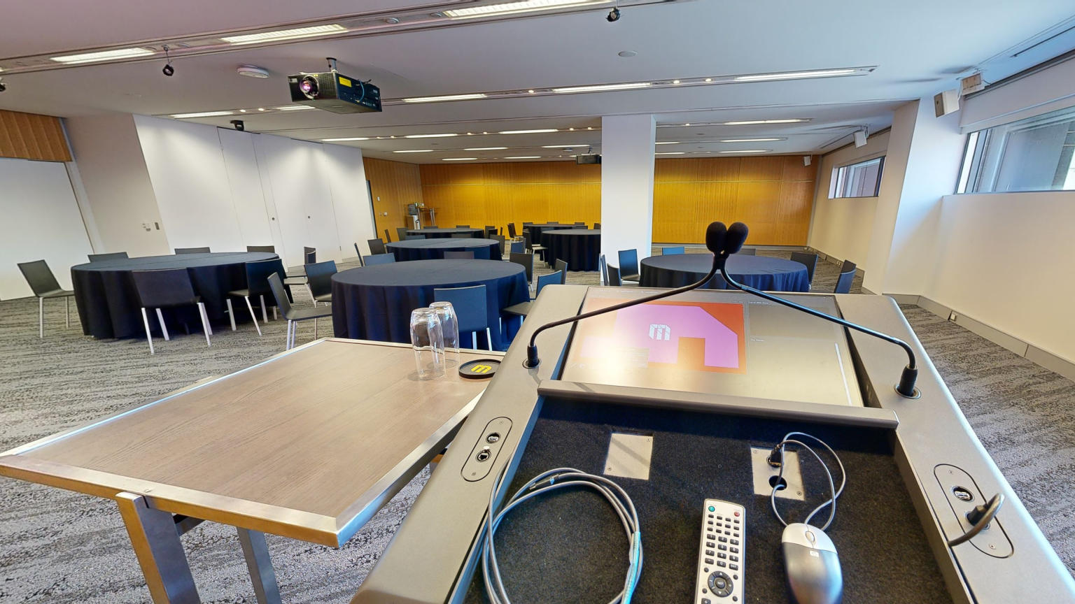 A conference or meeting room with arranged round tables and chairs for meetings or discussions. The tables focus around facing a lectern at the front of the room. A strip of windows that sit at eye level run along side one wall. 