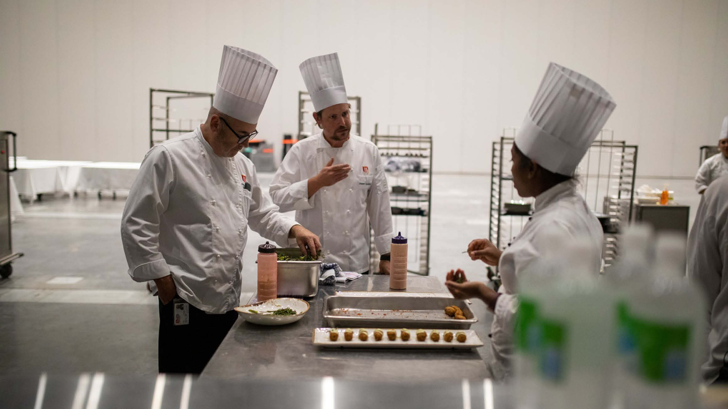 Three chefs in MCEC (Melbourne Convention and Exhibition Centre) uniform stand at an industrial kitchen bench in conversation.  