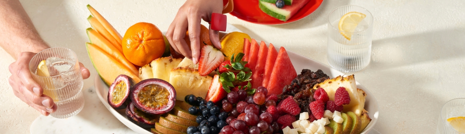 A large platter of fresh fruits cut up with a hand reaching over the plat to pick a strawberry. 