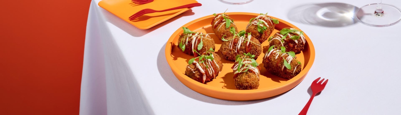 Potato bravas croquette with spicy tomato ketchup and aioli presented on an orange plate sitting on a white table cloth. 