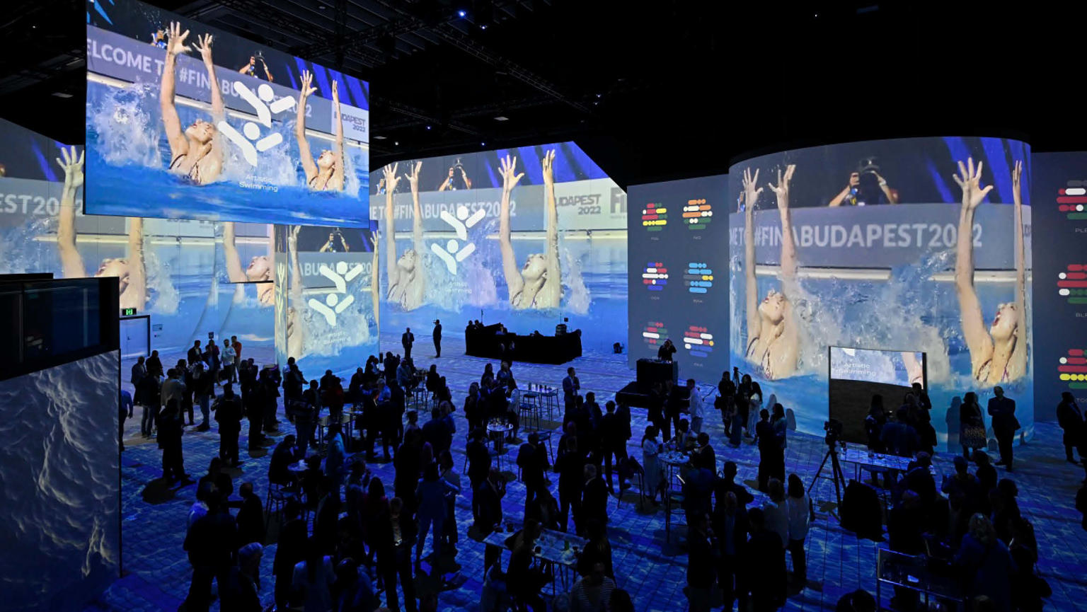 A large crowd assembled in a room enveloped by expansive digital screens forming the walls. The screens showcase captivating visuals of swimmers in a swimming pool.