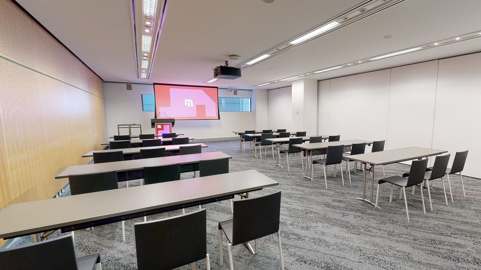 A meeting or conference room featuring neatly arranged tables and chairs in rows, providing a professional setting for meetings and discussions. The tables and chairs face a large projector screen and lectern at the front of the room. A small strip of windows that sit at eye level run behind the screen. 