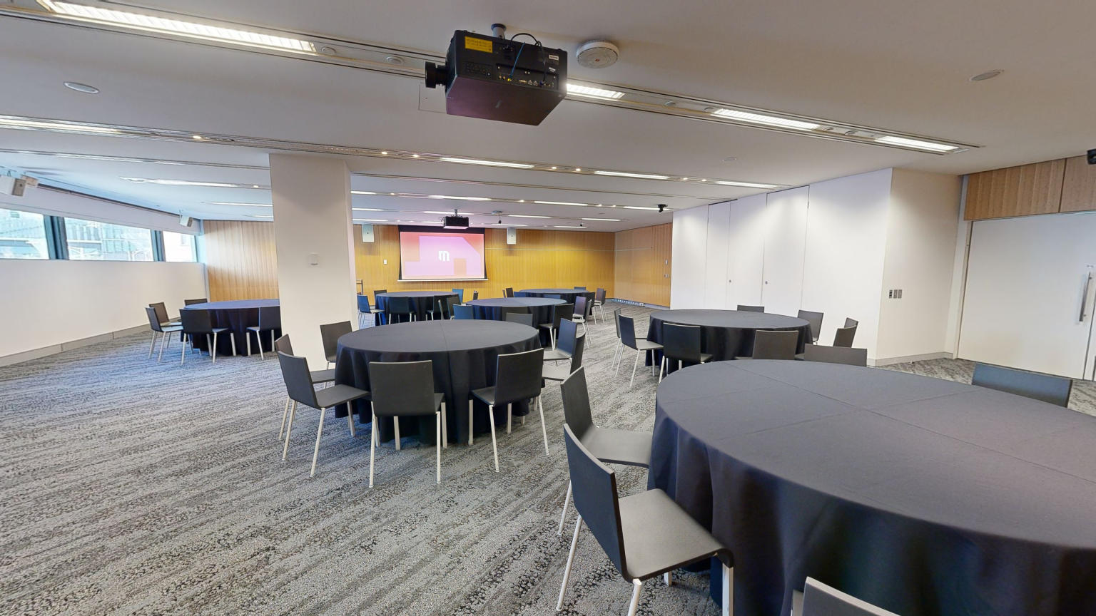 A conference or meeting room with arranged round tables and chairs for meetings or discussions. The tables focus around facing a large projector screen at the front of the room with a lectern to the left side. A strip of windows that sit at eye level run along side the far wall. 