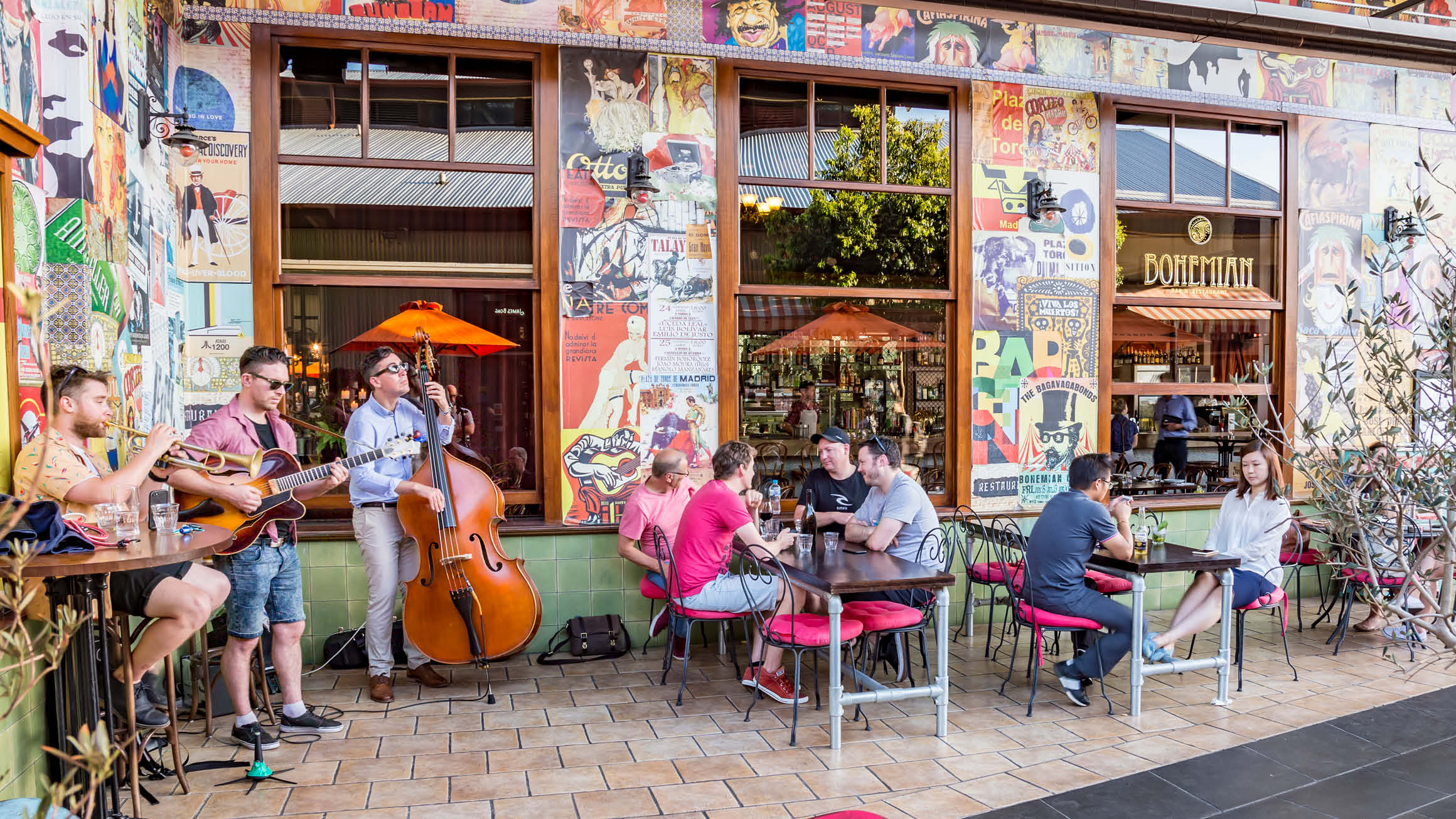 An inviting outdoor setting where people are enjoying conversation and relaxation outside a beautifully decorated restaurant. In the background, two talented musicians captivate the scene—one playing a guitar and the other a double bass guitar.