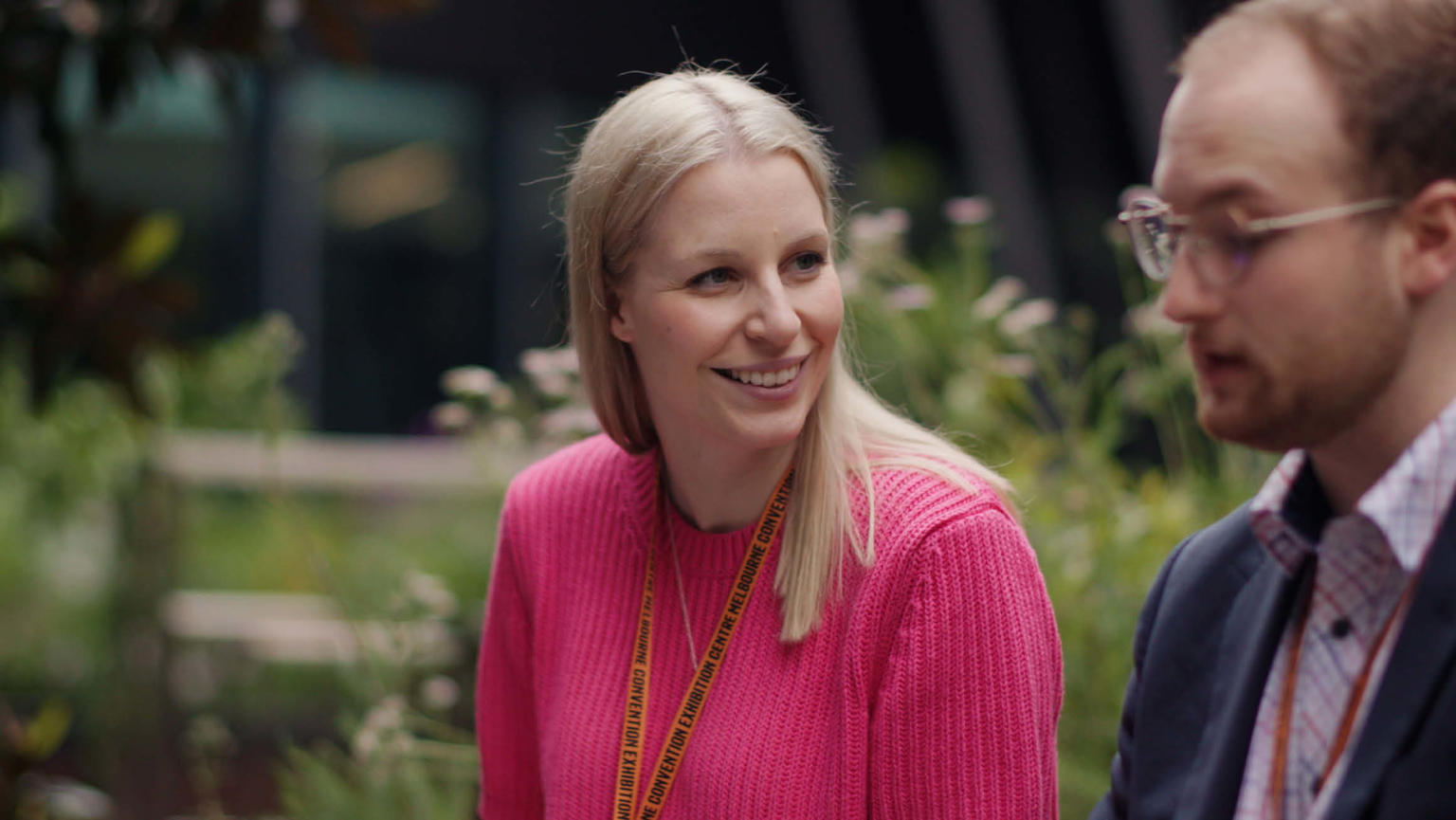 Anthea Fahey, wearing a pink jumper, sitting outdoors next to a man in a suit and glasses