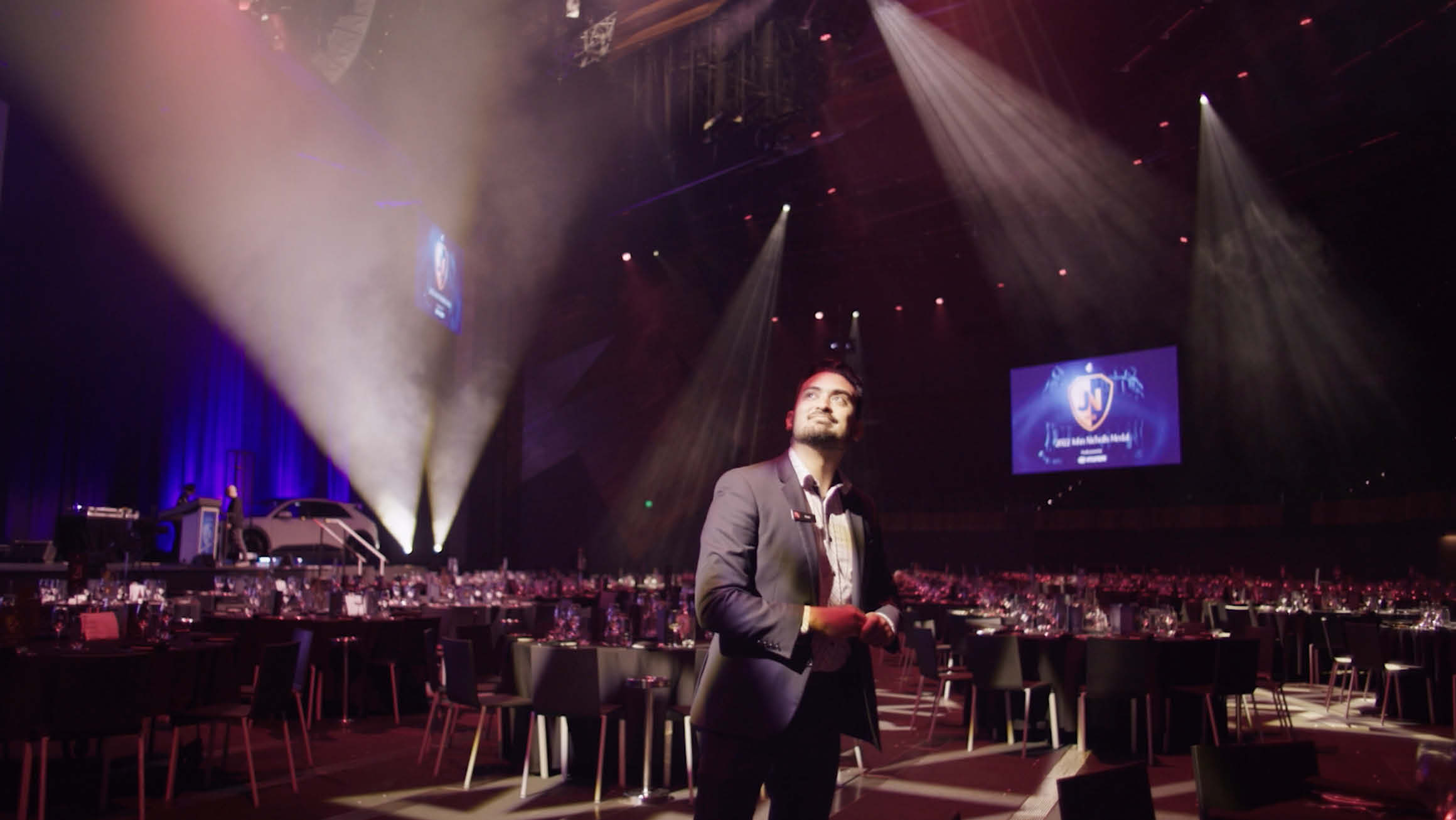 A man dressed in a suit stands in a spacious event room adorned with round tables and chairs. The room is illuminated with dynamic strobe lighting, and a captivating digital screen.