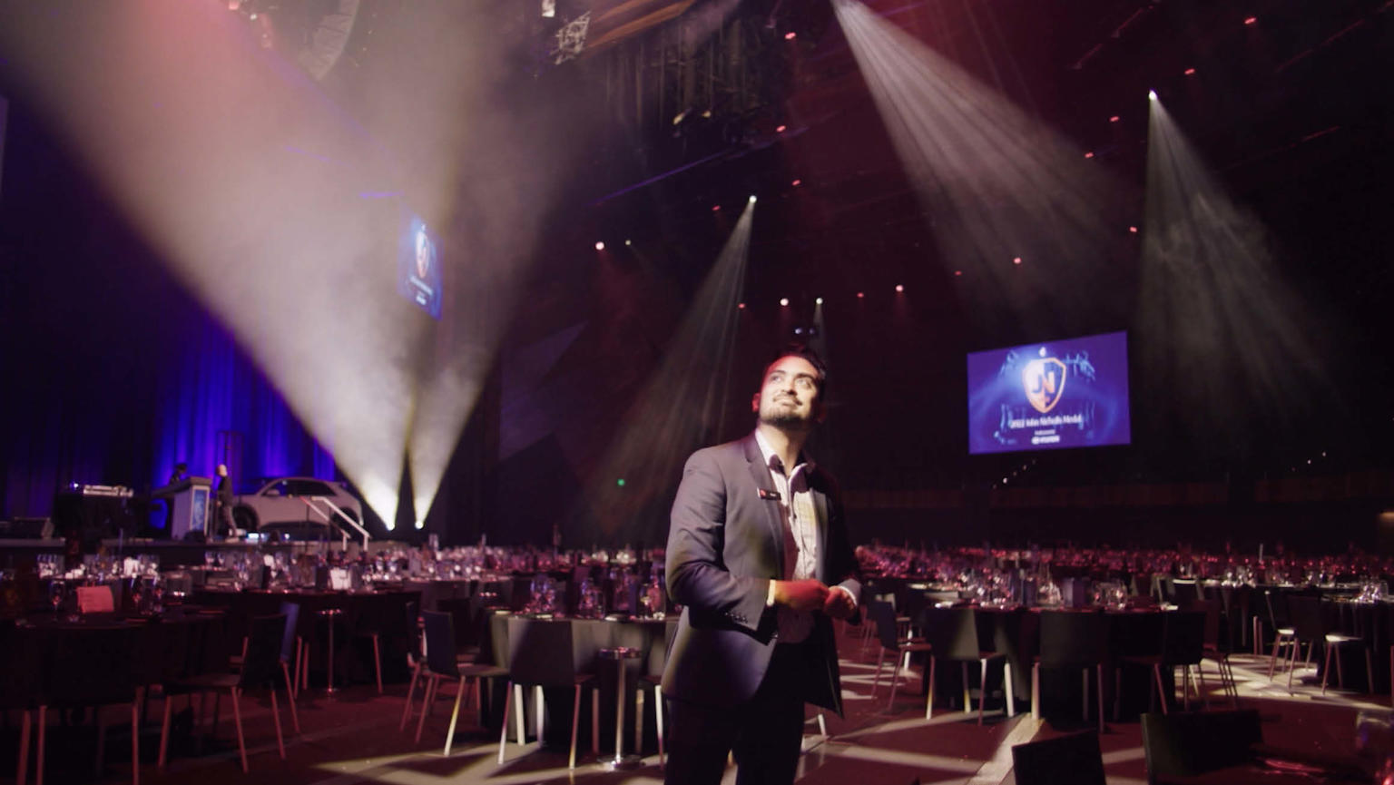 A man dressed in a suit stands in a spacious event room adorned with round tables and chairs. The room is illuminated with dynamic strobe lighting, and a captivating digital screen.