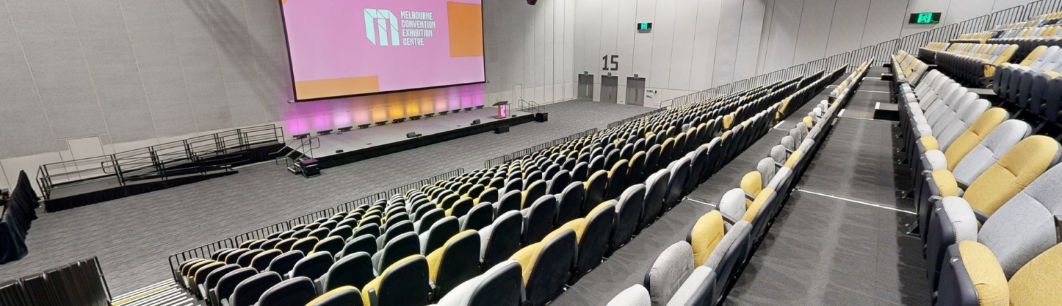 Empty theatre or yellow and grey chairs facing a giant screen at the front of the room. The screen is displaying a white MCEC logo on a pink and red background.