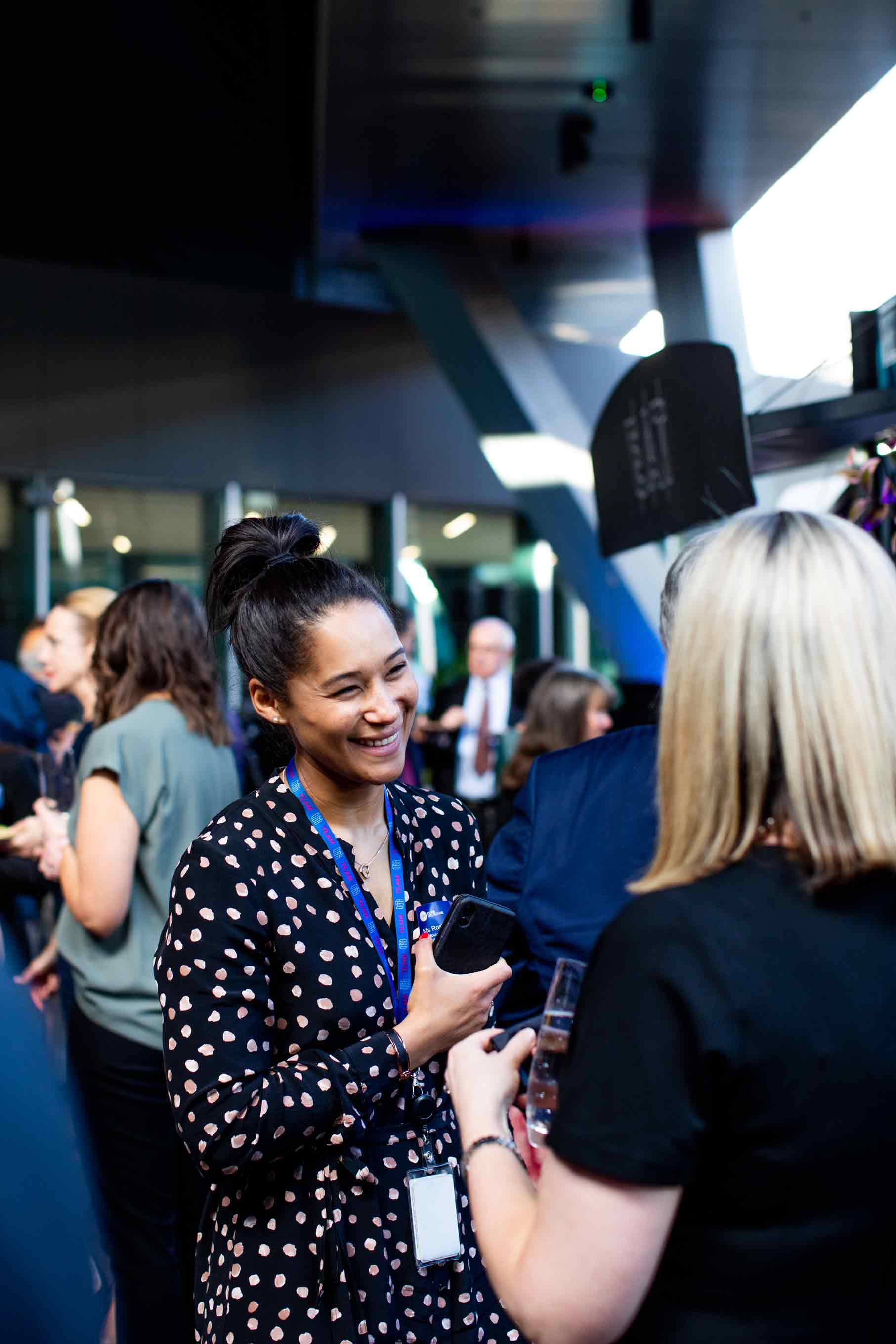 A woman wearing a black and white spotted dress laughs while talking to blonde lady wearing a black dress with her back towards the camera. They stand amongst a crowd of people. 