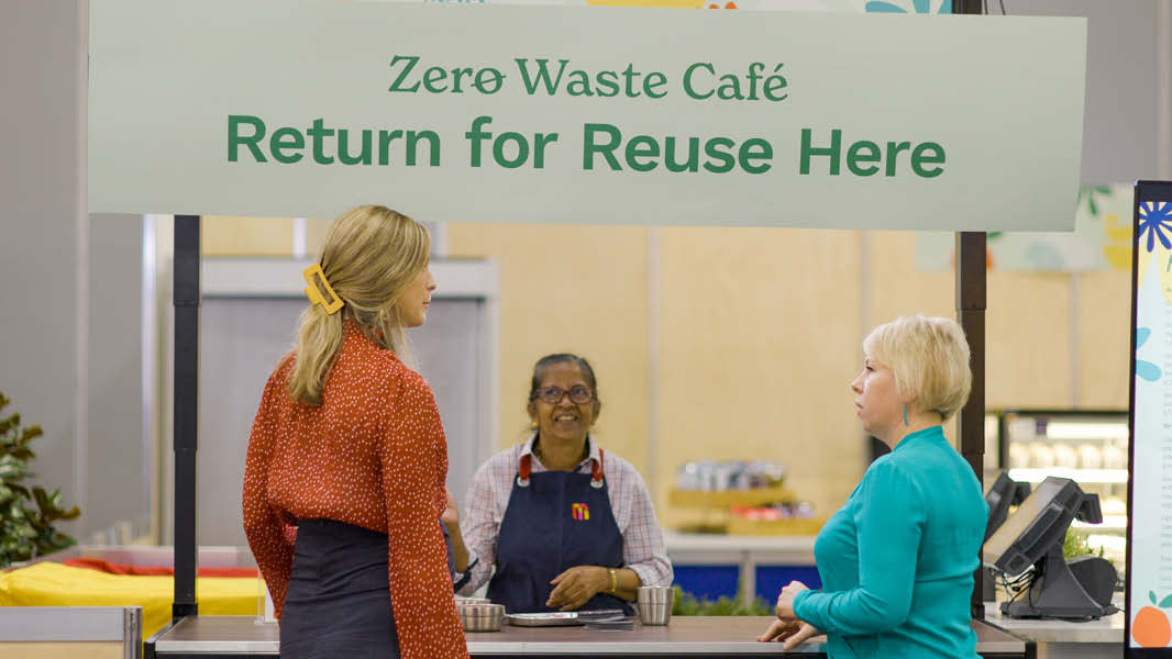 Two women, one in an orange jumper and the other in a teal top are talking to another women standing behind an exhibition stall with the word "Zero Waste Cafe, Return for Reuse Here" written on the display board over head. 