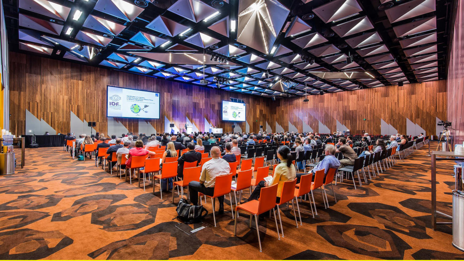 A gathering of individuals seated in neatly arranged rows of vibrant orange chairs, facing two prominent digital screens positioned at the front of the room. The walls exhibit a warm wooden texture, complemented by a patterned carpet.