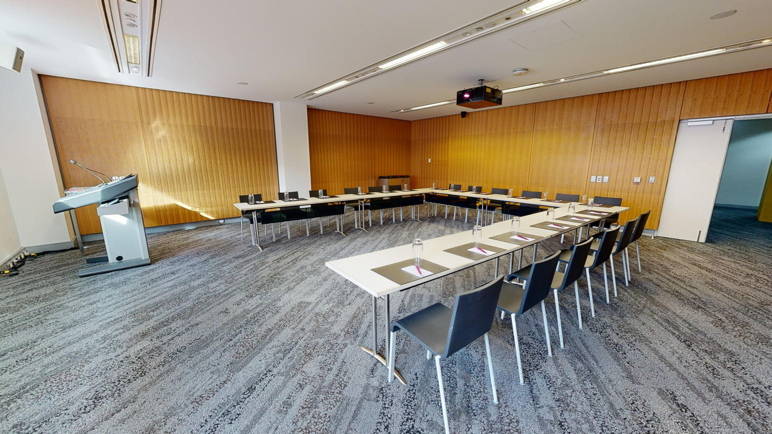 A meeting or conference room with tables and chairs arranged in a u-shaped boardroom configuration. The tables and chairs are facing a and lectern.