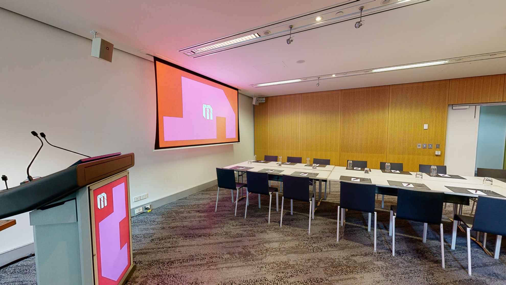 A conference or meeting room featuring a spacious boardroom layout equipped with a sizable screen and comfortable chairs for attendees. A lectern also sits in the corner to the left of the screen.