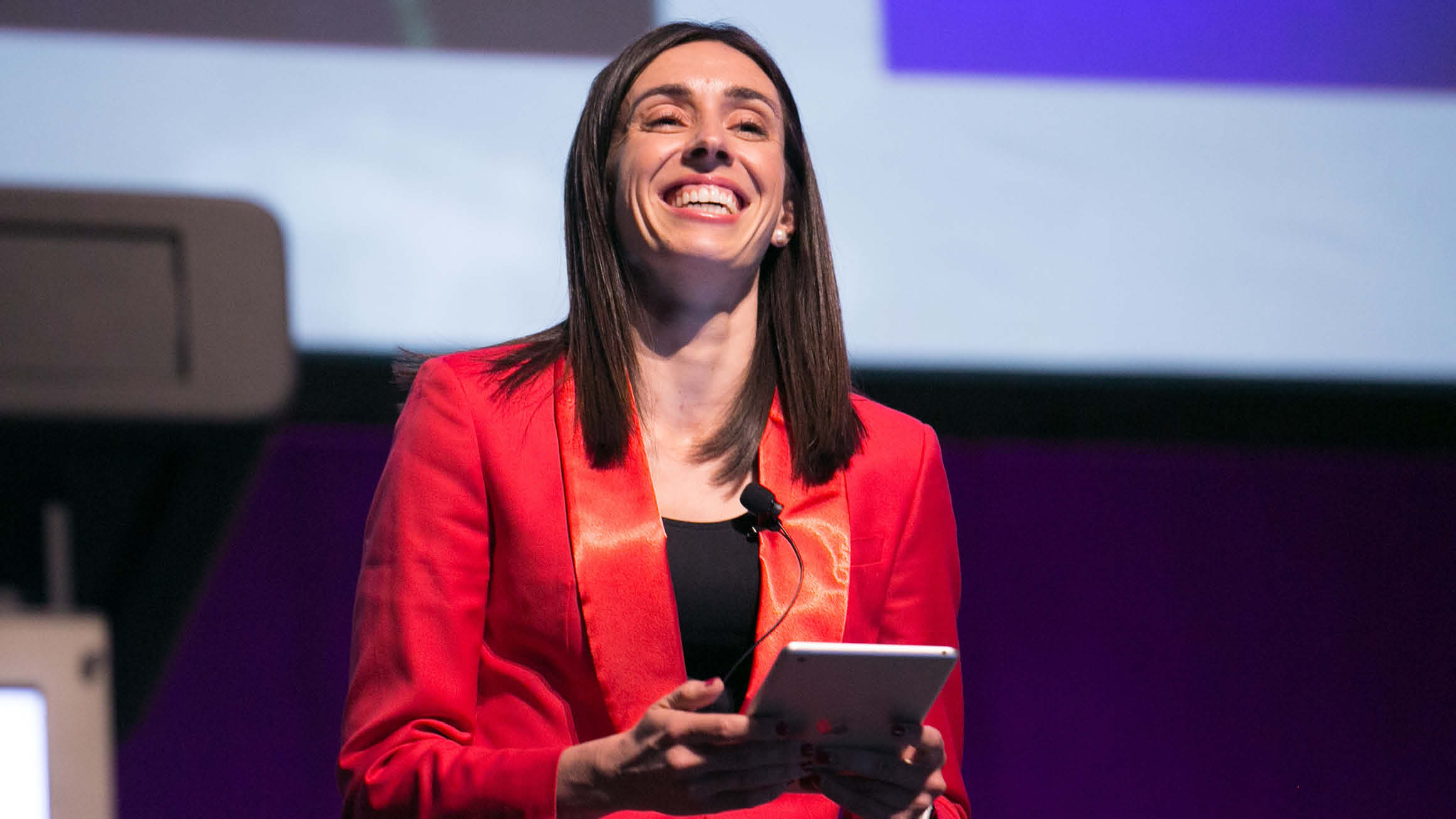 A confident woman wearing a vibrant red blazer stands at a podium, holding a tablet and addressing a crowd with a smile. The focus of her attention is behind the camera, creating an engaging and energetic atmosphere.