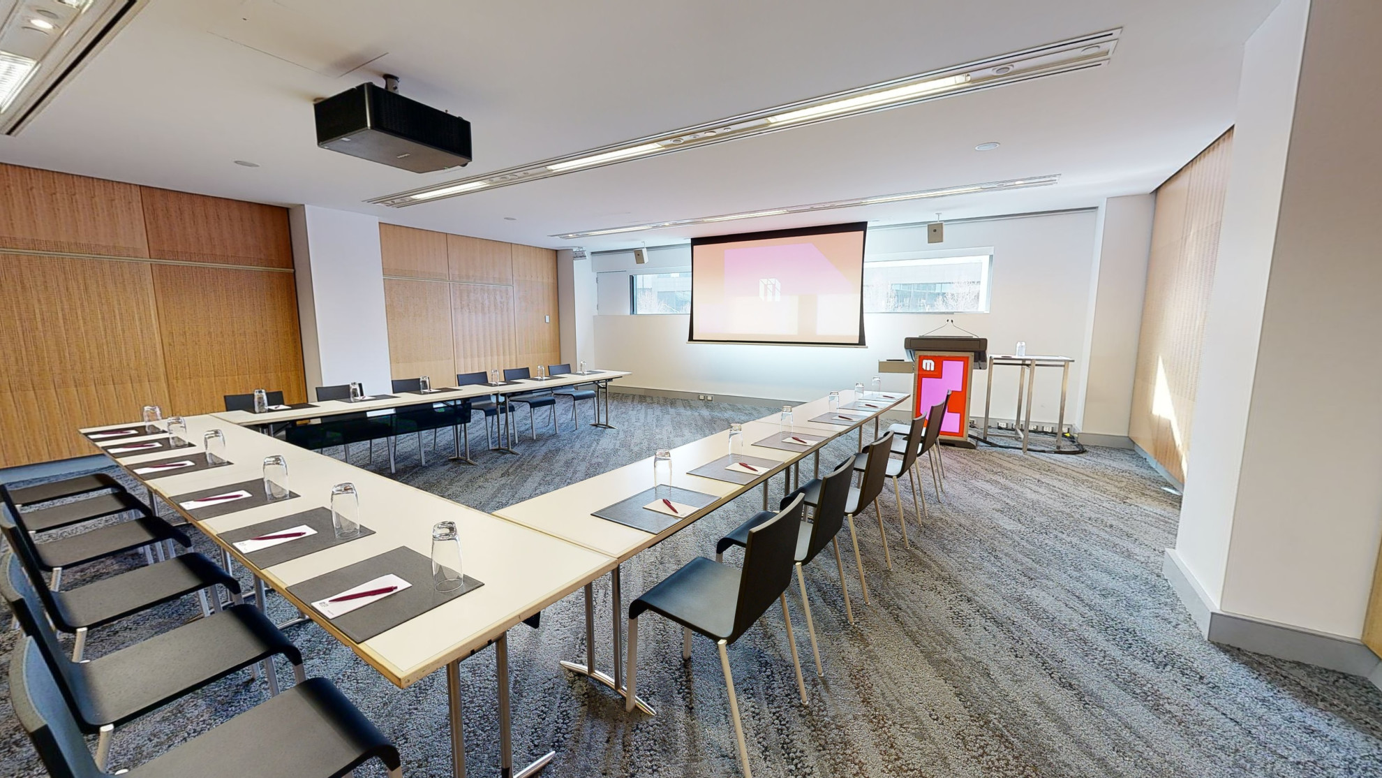 A meeting or conference room with tables and chairs arranged in a u-shaped boardroom configuration. The tables and chairs are facing a large projector screen and lectern both which have MCEC branding displayed on it. 