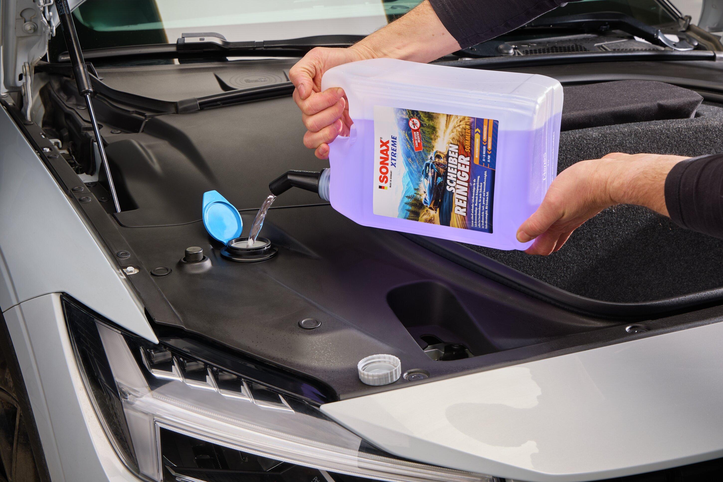 Jemand füllt den XTREME Schei­ben­Rei­niger aus dem 5-Liter-Kanister in den geöffneten Wischwassertank im Motorraum eines Autos. 